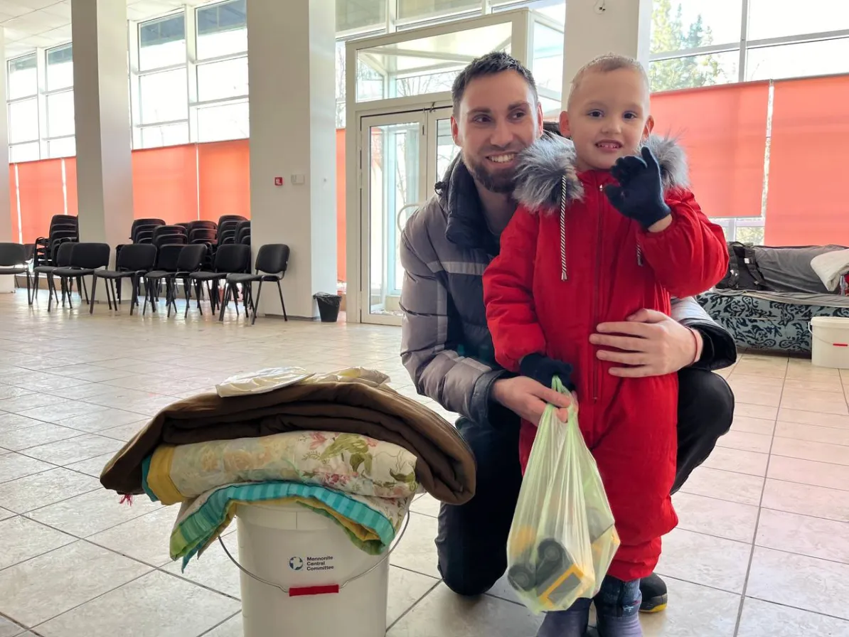 A man and a child pose together, smiling. The child wears a coat, and there are blankets stacked on a bucket nearby. Chairs are in the background.