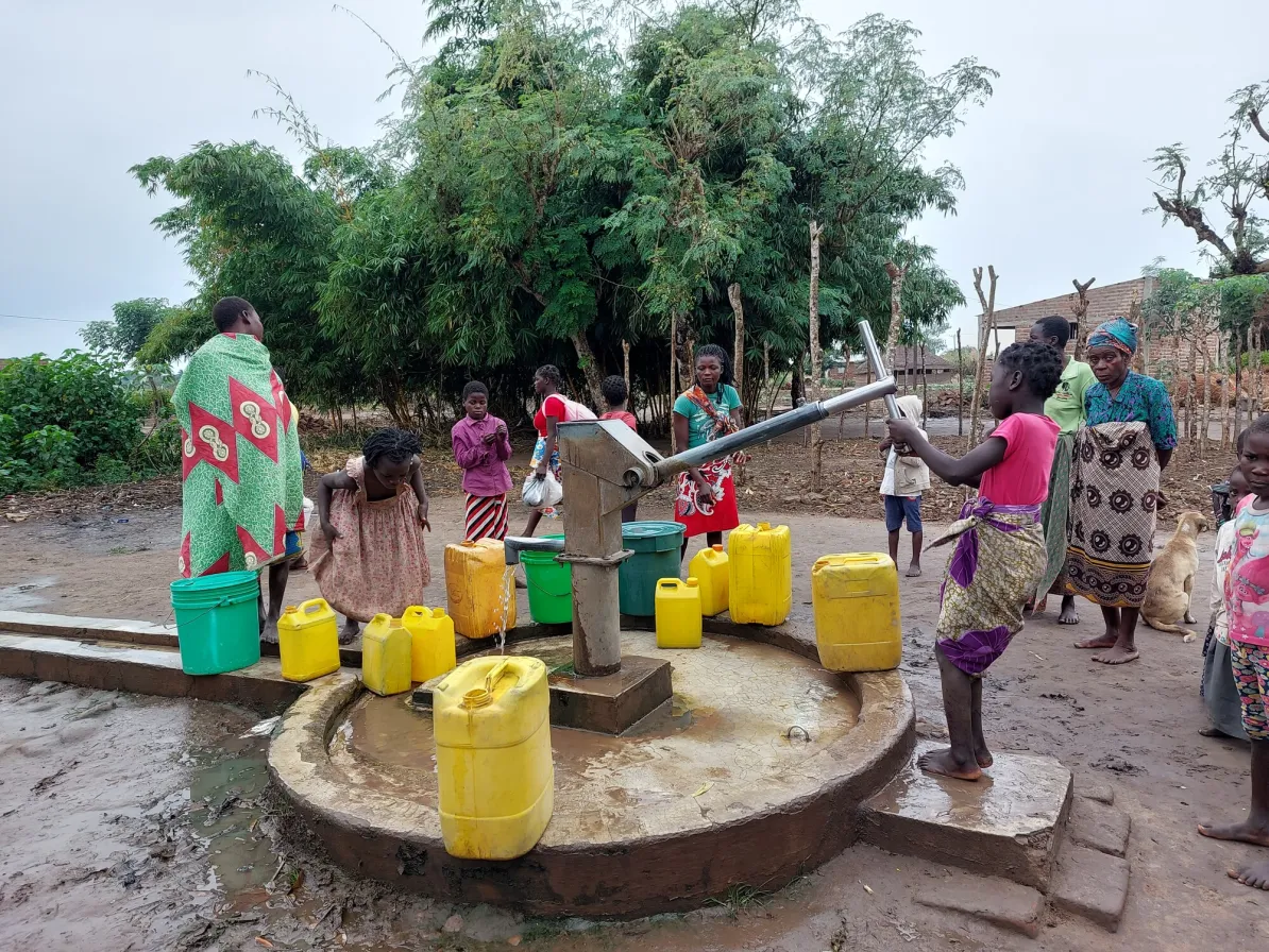 People are gathered around a water pump in a rural setting, filling yellow containers with water. Vegetation and overcast skies are visible in the background.