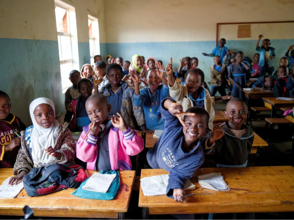 A classroom filled with children, some smiling and raising their hands, seated at wooden desks with books open in front of them.