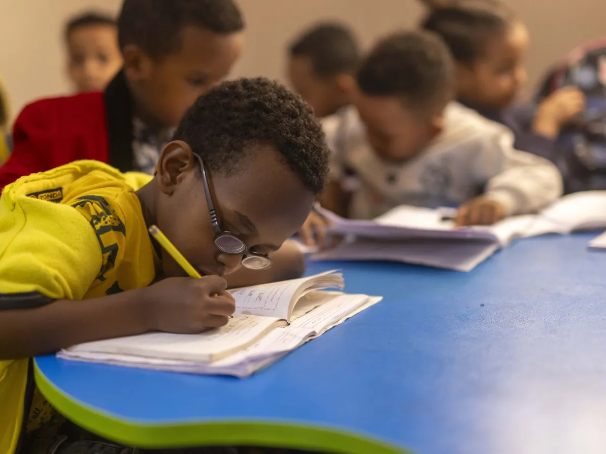 A group of children is seated at a table, focused on writing in notebooks. One child wears glasses and leans over a book.