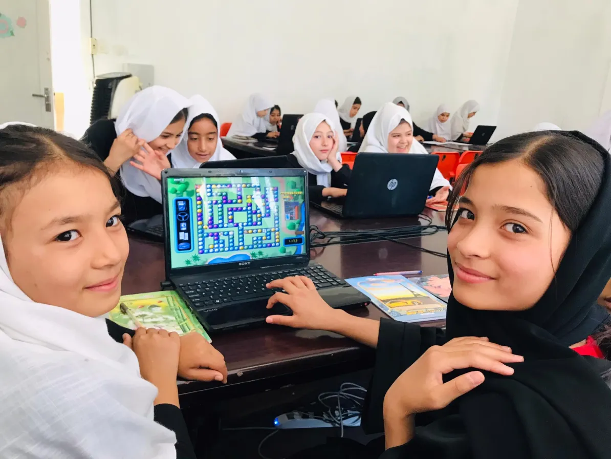 A group of girls in headscarves sit at desks with laptops, engaged in a computer activity, smiling and interacting with each other.