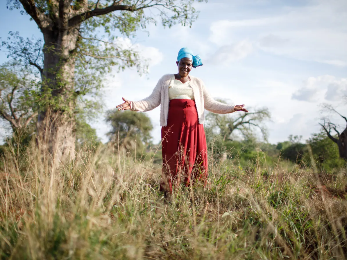 A woman stands in a grassy field, arms outstretched, with trees and a cloudy sky in the background.