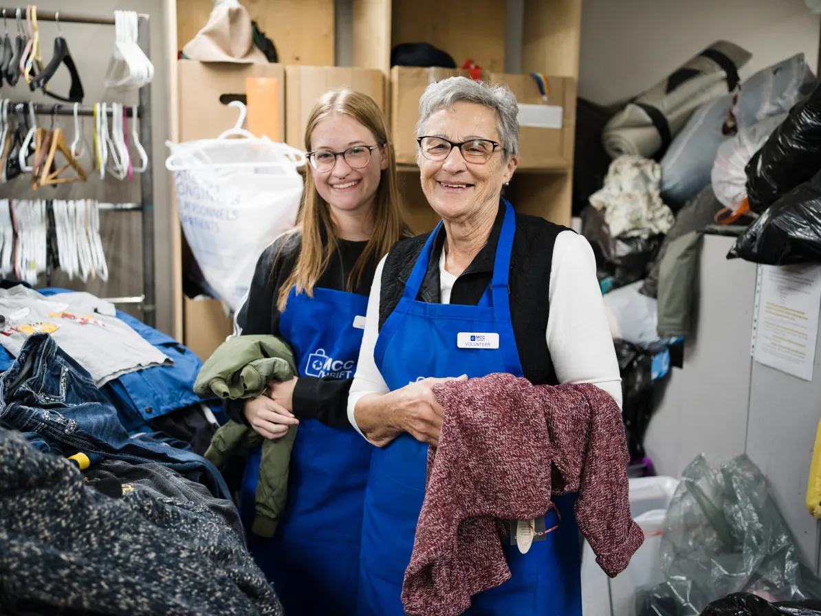 Two smiling women wearing blue aprons take a break sorting through clothing donations at an MCC Thrift Shop.