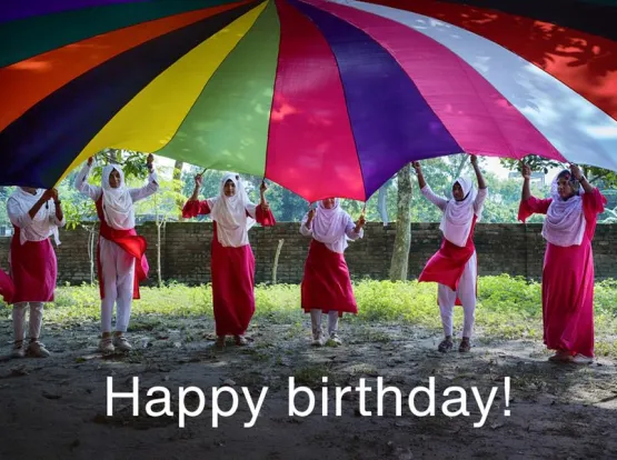 A group of girls lifting a colorful parachute overhead and the words "Happy birthday!"