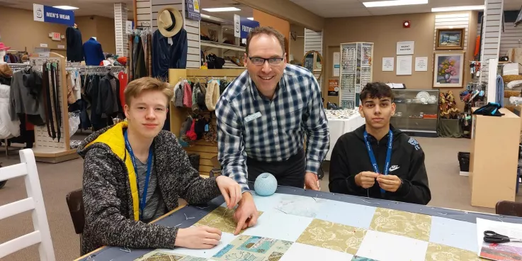 Two youth and an adult pose for the camera while sitting around a comforter in a thrift shop.
