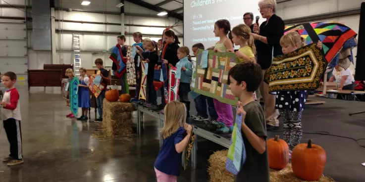 Several children stand on a stage holding up quilted wall hangings. There is an older woman behind them speaking into a microphone. 
