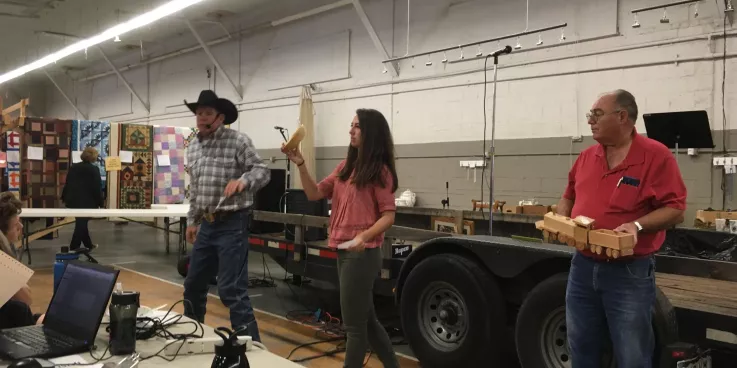 An auctioneer in a black cow boy hats stands next to a woman holding auction item, a wooden bowl. Next to here is a man holding another auction item, a wooden toy train.