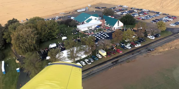 An overview view of a church building during an event. There are a lot of cars parked around the church. The photo was taken out the window of a yellow airplane.