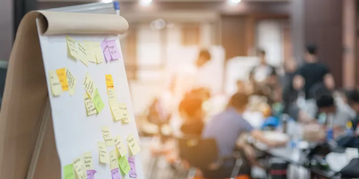 A flip chart board is in the foreground with sticky notes attached, while a group sits in the background.
