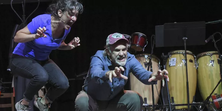 A man and a woman act on stage. The woman is jumping in the air, and the man is crouching.