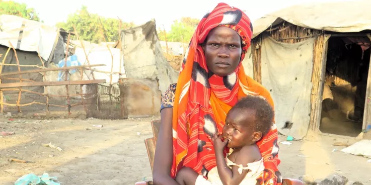 A woman holding a baby in front of a camp for internally displaced people.