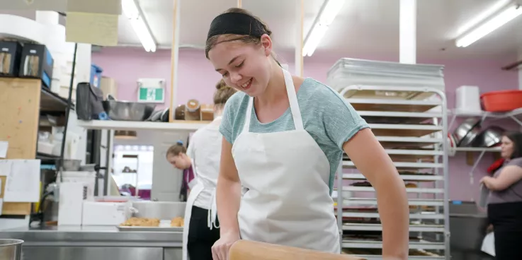 A young lady with a white apron uses a rolling pin to roll out dough.