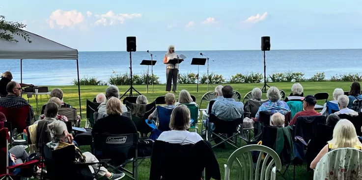 A woman speaks to a crowd of people in chairs outdoors with a lake in the background.