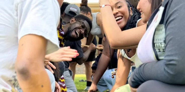 A group of smiling young people participate in a team-building activity that involves a stick on the ground.