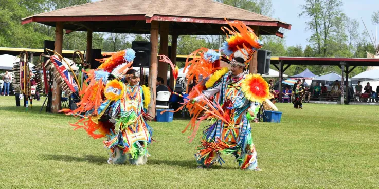 A father and son dance in colourful traditional Indigenous dress in front of a pavillion on green grass.