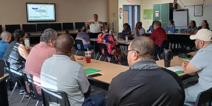 group of people sitting around a few tables listening to the main facilitator standing in the center