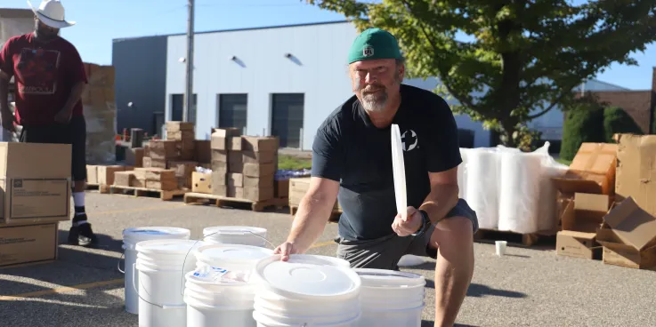 A man with a cap is kneeling next to several white buckets. Boxes are in the background.