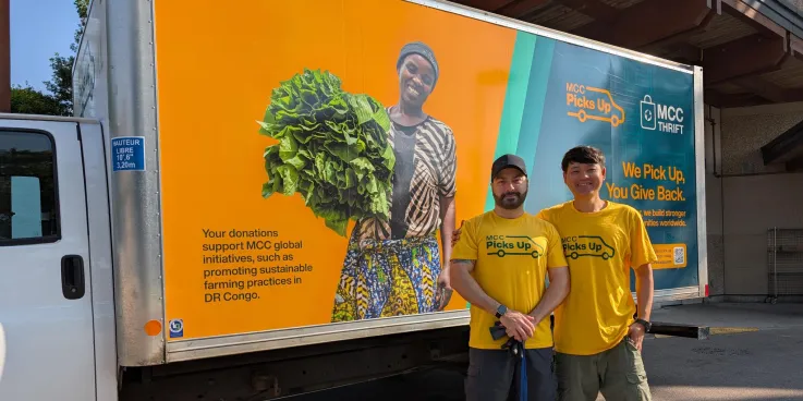Two men in yellow stand in front of an MCC Thrift truck that is also yellow with images on it.