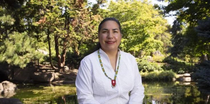 A woman wearing a white shirt sits in front of a lush green landscape.