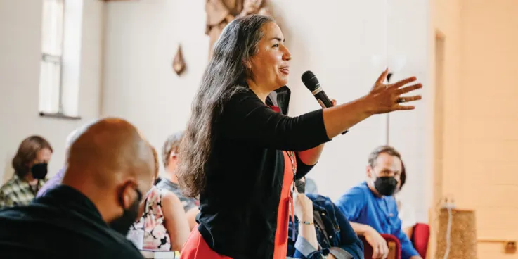 Sarah Augustine stands and holds a microphone while talking to a crowd of people sitting down.