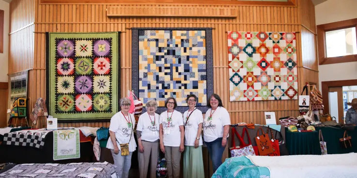 Five members of the Piece It Together quilting group stand in front of three quilts during an annual sale