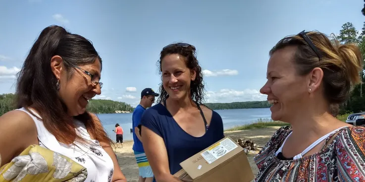 Three women holding boxes and comforters while standing near a shoreline.