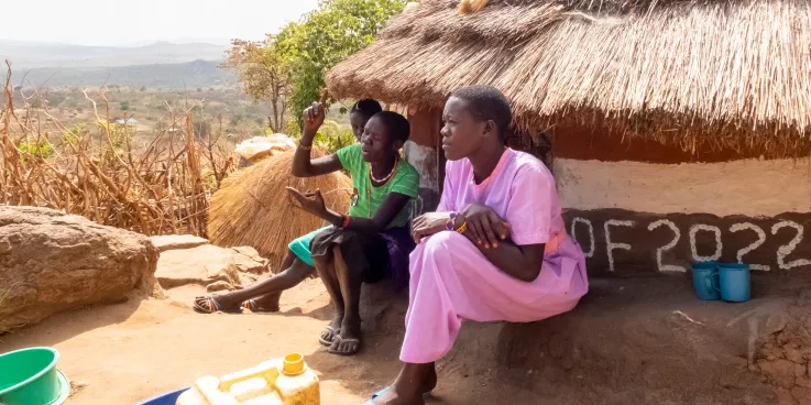 Three women sit in front of a hut home in Uganda.