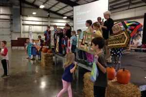 Several children stand on a stage holding up quilted wall hangings. There is an older woman behind them speaking into a microphone. 