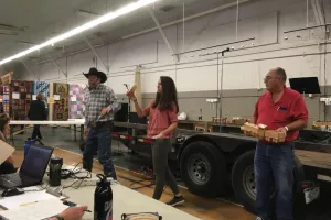 An auctioneer in a black cow boy hats stands next to a woman holding auction item, a wooden bowl. Next to here is a man holding another auction item, a wooden toy train.