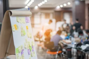 A flip chart board is in the foreground with sticky notes attached, while a group sits in the background.