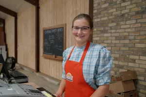 A young woman wearing a red apron stands behind a cash register at a country market.