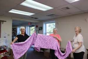 three women hold up a piece of pink fabric