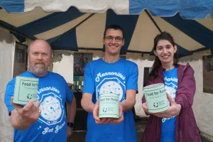 Standing outside a tent, three people wearing identical blue shirts each hold out a can that reads "Food for Relief" to the camera.