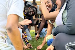 A group of smiling young people participate in a team-building activity that involves a stick on the ground.