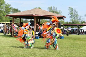 A father and son dance in colourful traditional Indigenous dress in front of a pavillion on green grass.
