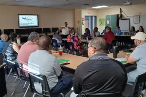 group of people sitting around a few tables listening to the main facilitator standing in the center