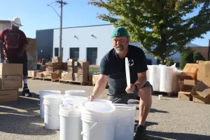 A man with a cap is kneeling next to several white buckets. Boxes are in the background.
