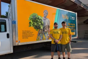 Two men in yellow stand in front of an MCC Thrift truck that is also yellow with images on it.