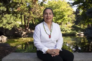 A woman wearing a white shirt sits in front of a lush green landscape.