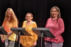 Three women stand behind music stands in a dark room.