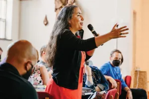 Sarah Augustine stands and holds a microphone while talking to a crowd of people sitting down.