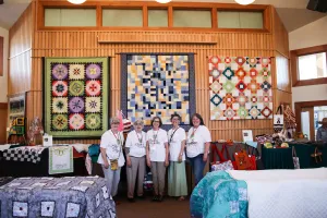 Five members of the Piece It Together quilting group stand in front of three quilts during an annual sale