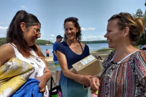 Three women holding boxes and comforters while standing near a shoreline.