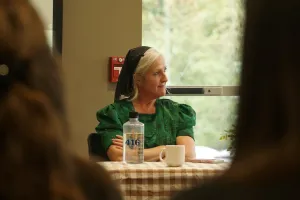 A woman dressed in traditional Mennonite clothing and head cover sits at a table in conversation.