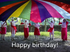 A group of girls lifting a colorful parachute overhead and the words "Happy birthday!"