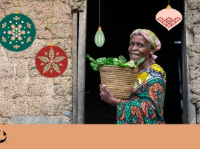 A woman holding a box of green vegetables. Christmans ornaments are imposed over the photo with a MCC logo in the bottom left corner.