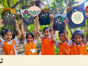 Young girls smiling holding up artwork. Christmas ornaments are overlaid on the photo with the MCC logo in the bottom left corner.