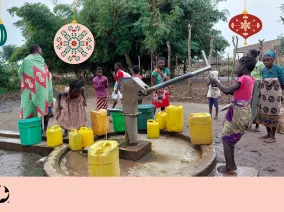 A group of children pumping water from a well. Christmas ornaments are imposed over the image with the MCC logo in the bottom left corner.