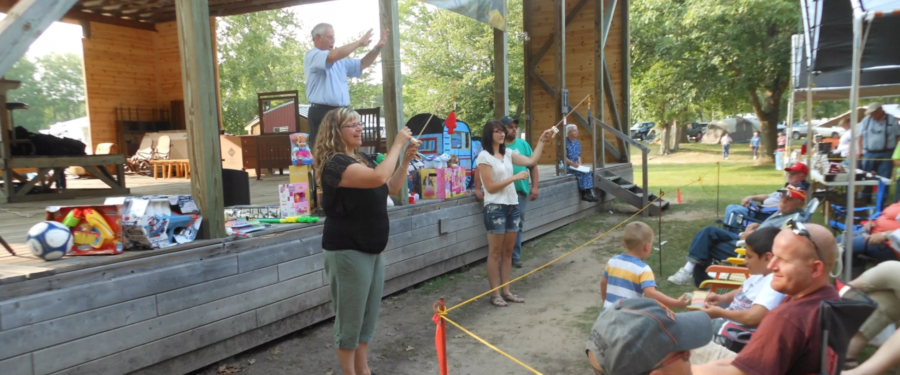 An auctioneer stands on an outdoor stage. Two women are down below displaying toys that are for auction.
