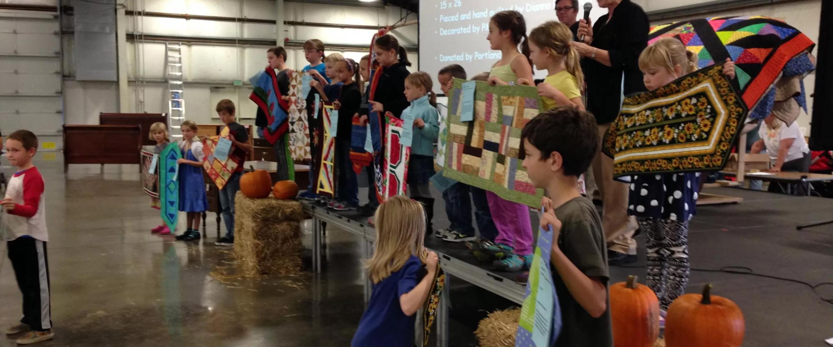 Several children stand on a stage holding up quilted wall hangings. There is an older woman behind them speaking into a microphone. 